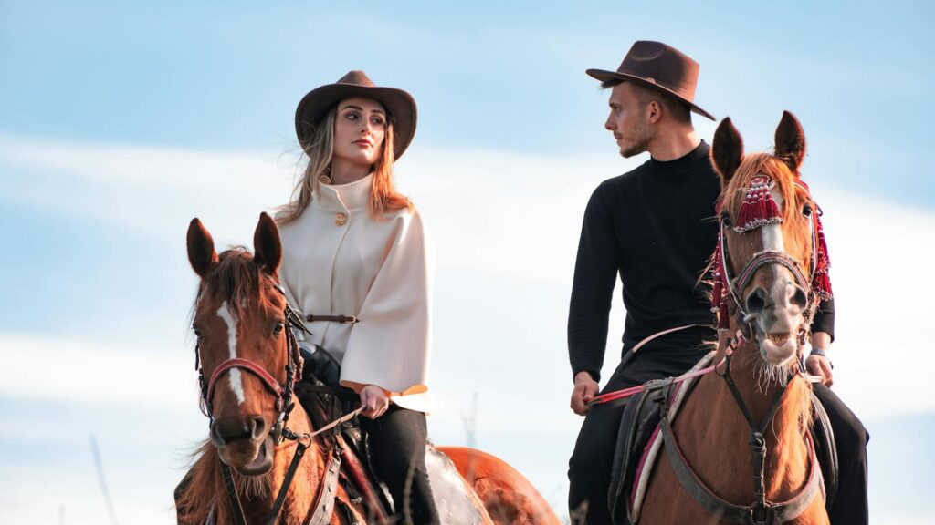 A man and woman in cowboy hats riding horses under blue sky, showcasing Western style outdoors.