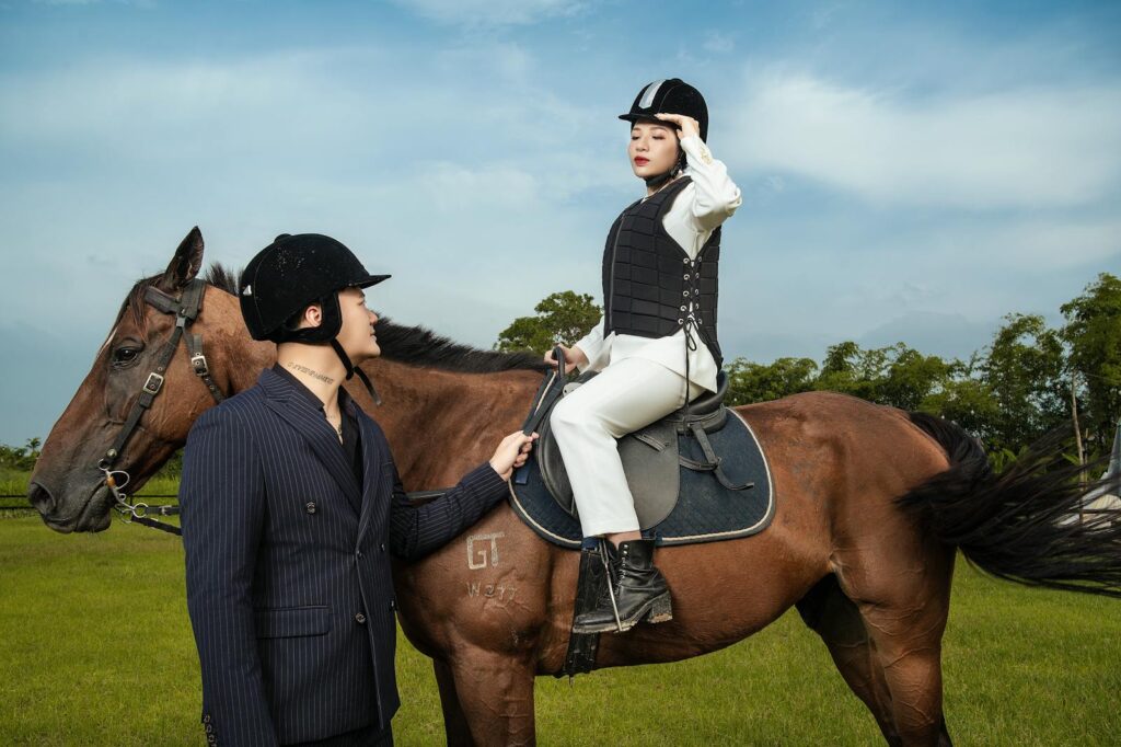 Two equestrians, one mounted and one standing, prepare a brown horse for a ride.