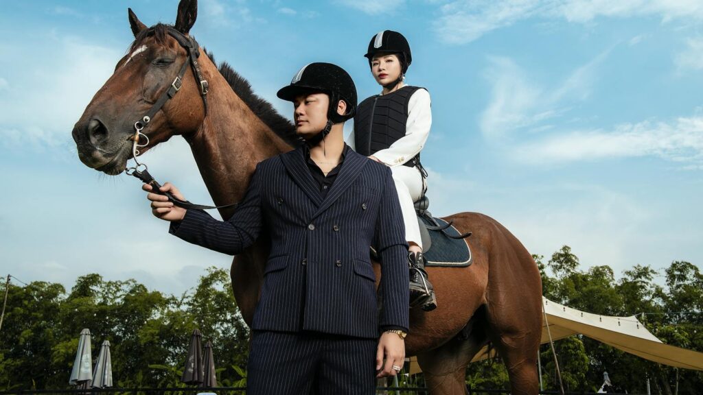 Two equestrian riders in helmets pose with a horse outdoors under a bright sky.