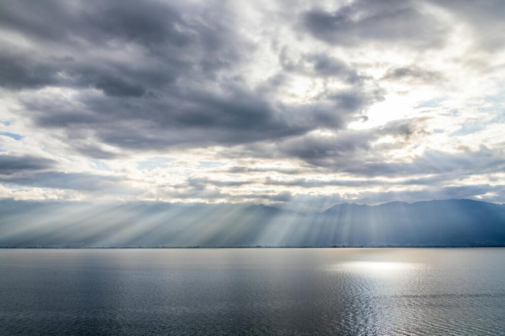 Beautiful sun rays pierce through clouds over a serene lake with mountains in the background.