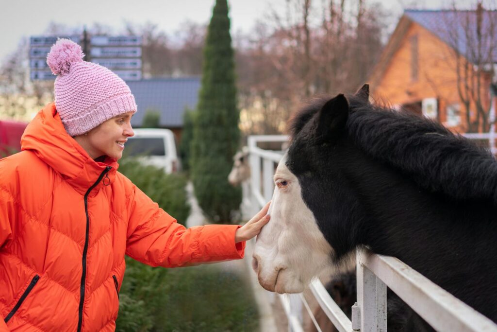 Smiling woman in a red jacket pets a horse on a farm during a winter day.