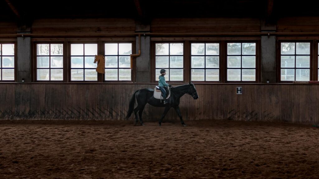 Girl riding a black horse indoors, showcasing equestrian training in a stable setting.