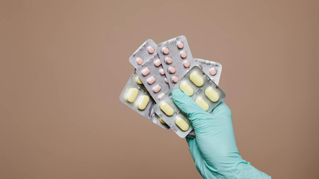 Close-up of a gloved hand holding multiple blister packs of tablets against a neutral background.