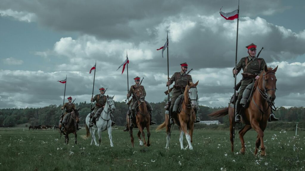 Five Polish cavalry soldiers on horses holding flags in an open field.