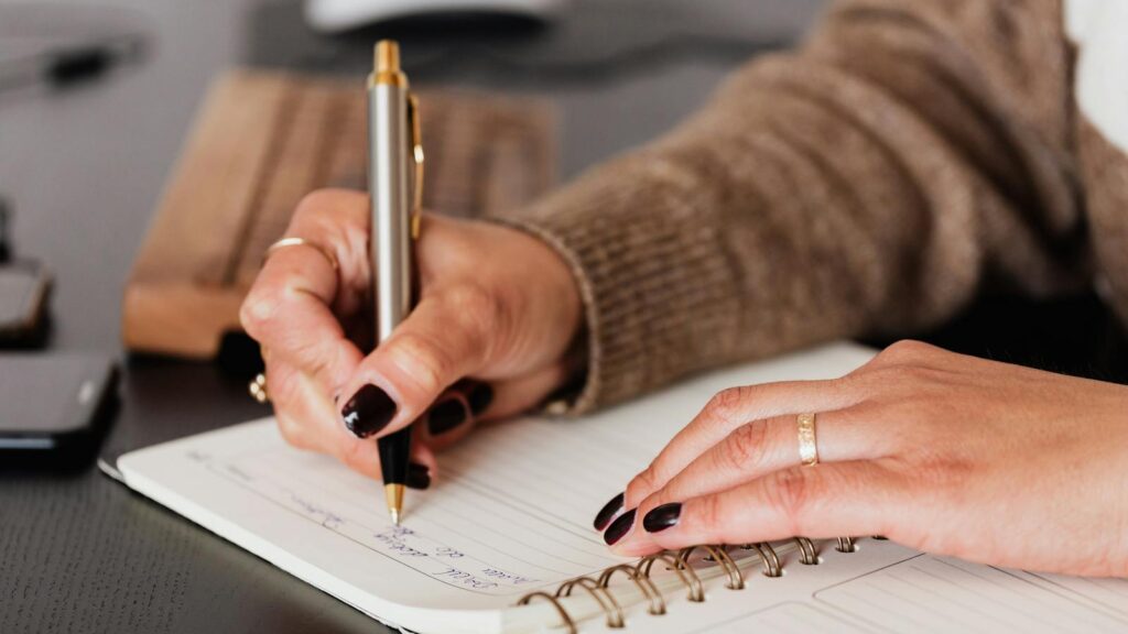 Crop unrecognizable female with stylish manicure sitting at black desk with keyboard and smartphone and taking notes with silver pen in notepad