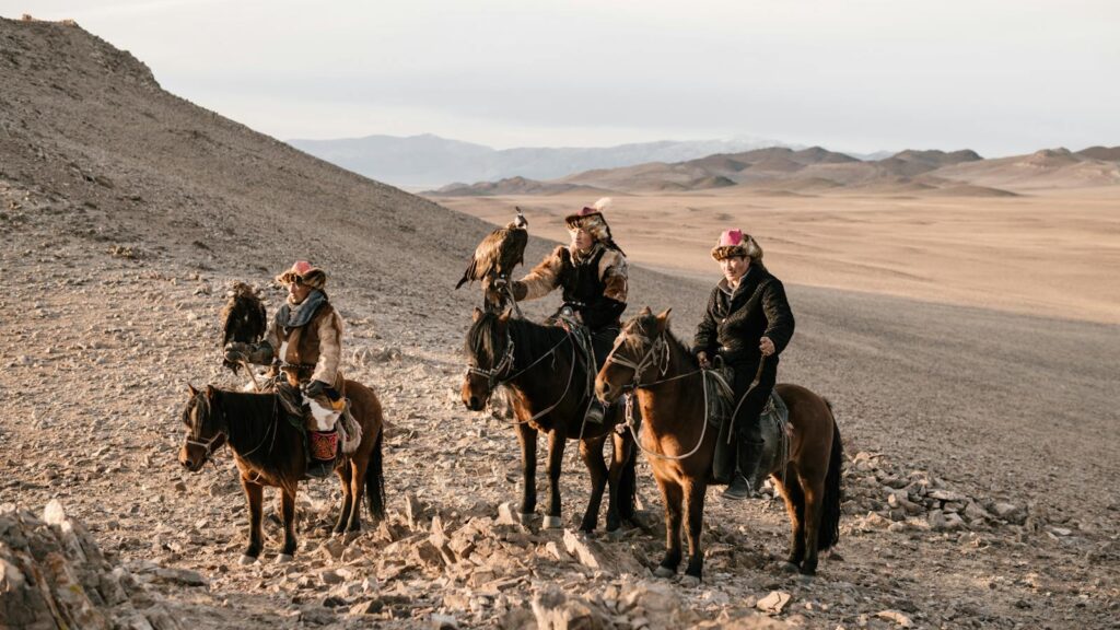 Three traditional eagle hunters riding horses in the arid Mongolian landscape, showcasing falconry skills.