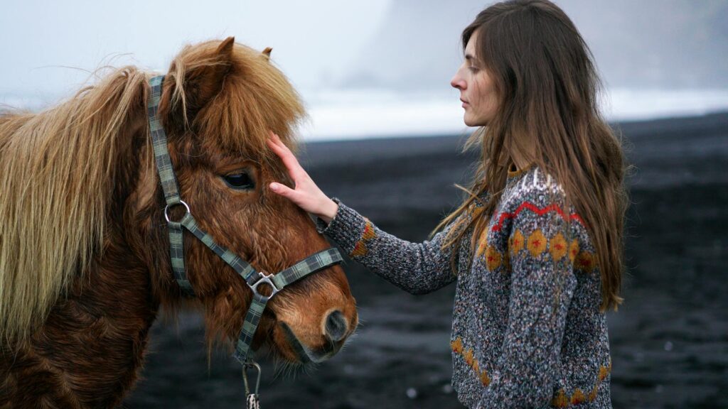 A woman gently pats a horse on a black sand beach, showcasing tranquility by the sea.