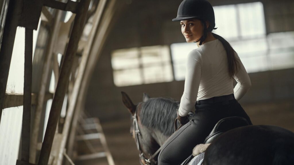 A woman elegantly rides a horse indoors in an equestrian setting, looking back with a smile.