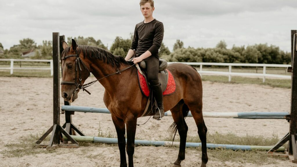 A young man sits confidently on a horse in an outdoor equestrian arena.