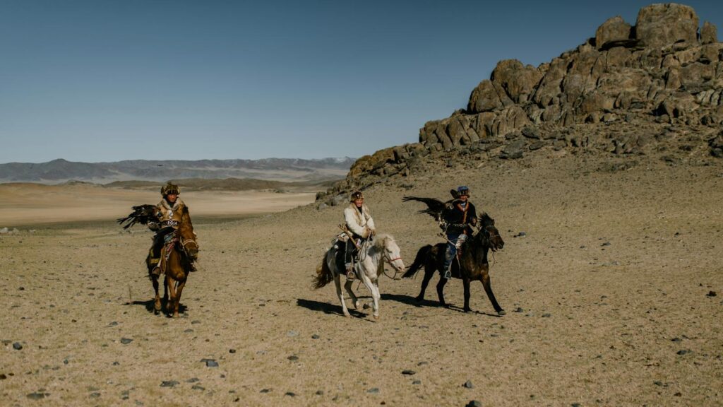 Three nomadic horse riders with eagles in a vast arid desert setting, showcasing traditional attire and horsemanship.