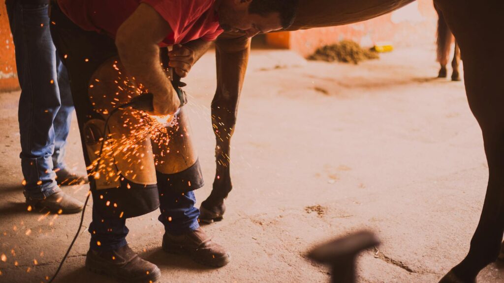 A farrier shoeing a horse in an indoor stable with flying sparks from the equipment.