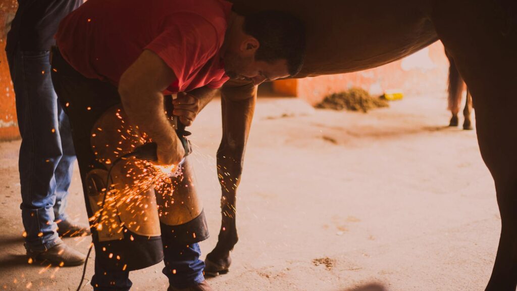 A farrier shoeing a horse in an indoor stable with flying sparks from the equipment.