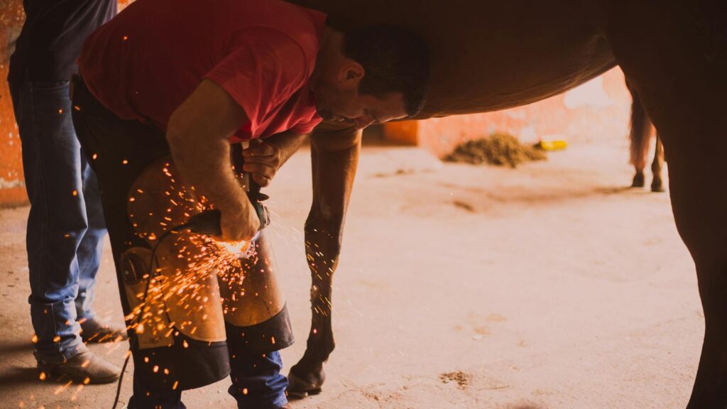 A farrier shoeing a horse in an indoor stable with flying sparks from the equipment.