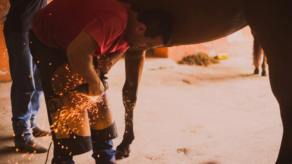 A farrier shoeing a horse in an indoor stable with flying sparks from the equipment.