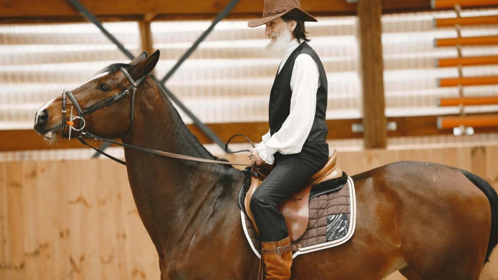 Elderly cowboy riding a brown horse indoors, side view, rustic setting.