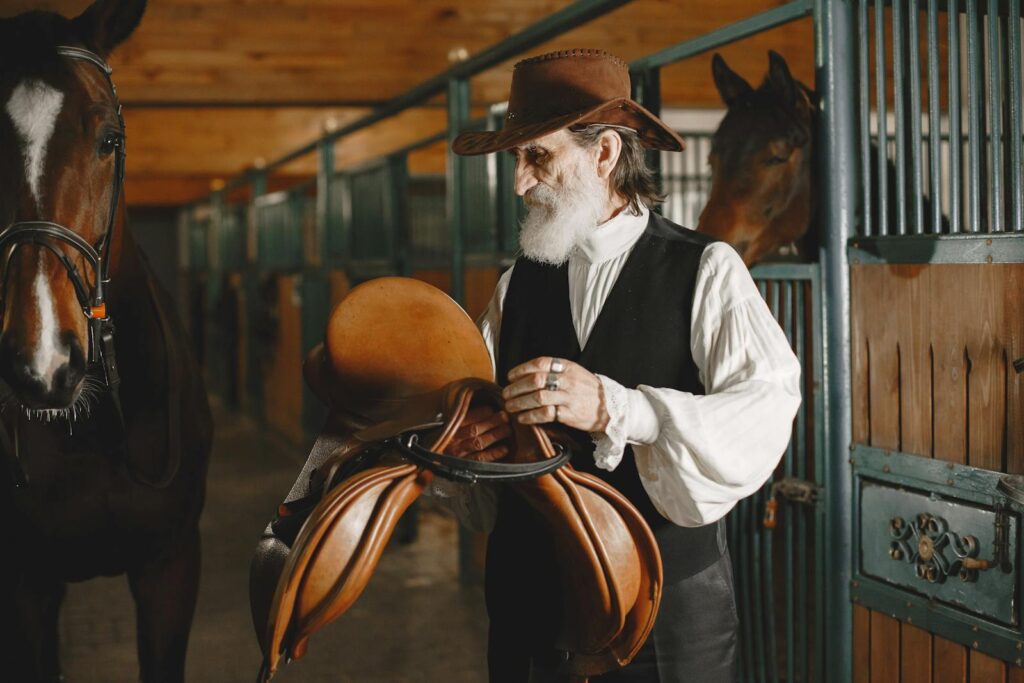 Elderly cowboy preparing saddle in stable with horses nearby