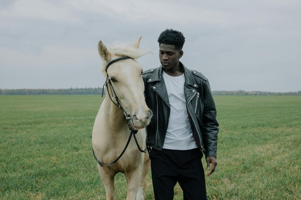 A man in a leather jacket stands beside a horse in an open grassland.