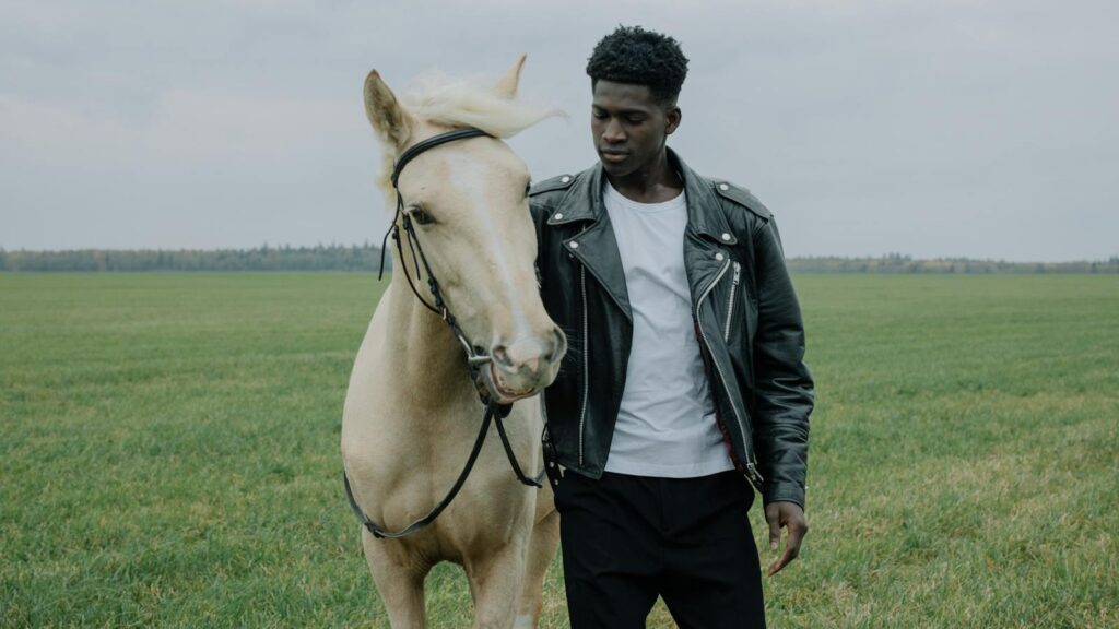A man in a leather jacket stands beside a horse in an open grassland.
