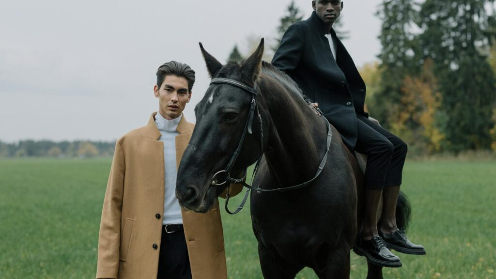 Two stylish models in elegant coats and a horse, set in a serene field.