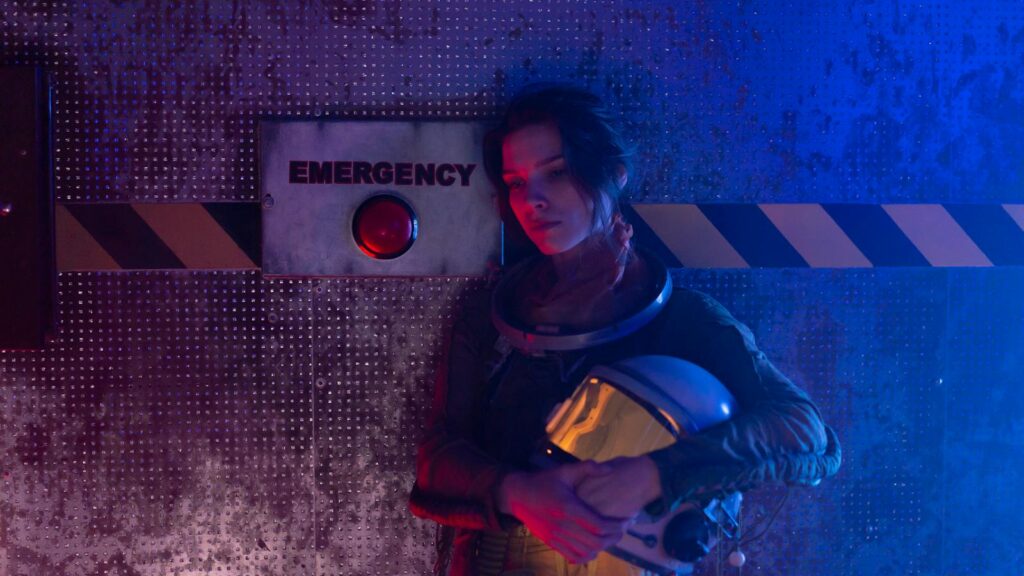 Young astronaut holding helmet in a dark emergency-themed room with red and blue lighting.