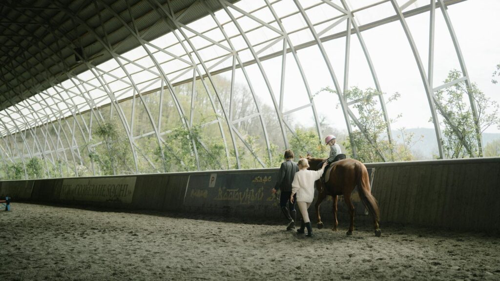 Child learning horseback riding in an indoor equestrian arena with guidance from instructors.