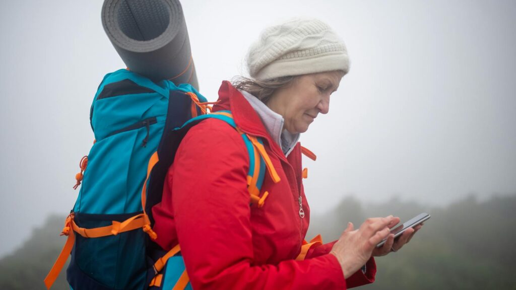 Elderly woman in red jacket checks phone while hiking in foggy scenery outdoors.