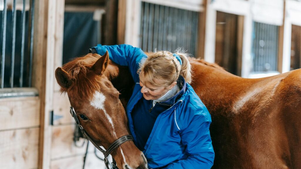 A woman lovingly embraces a horse in a rustic stable setting, showcasing equestrian care and bonding.