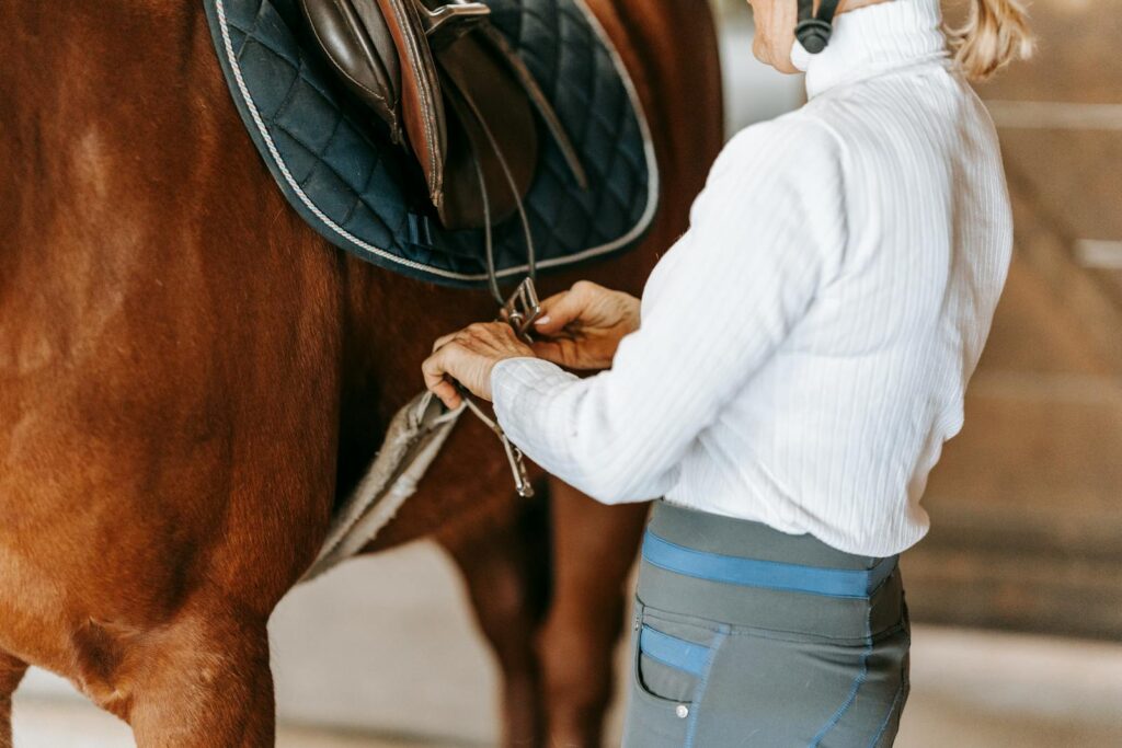 An equestrian woman adjusts a saddle on a horse inside a stable, preparing for a ride.