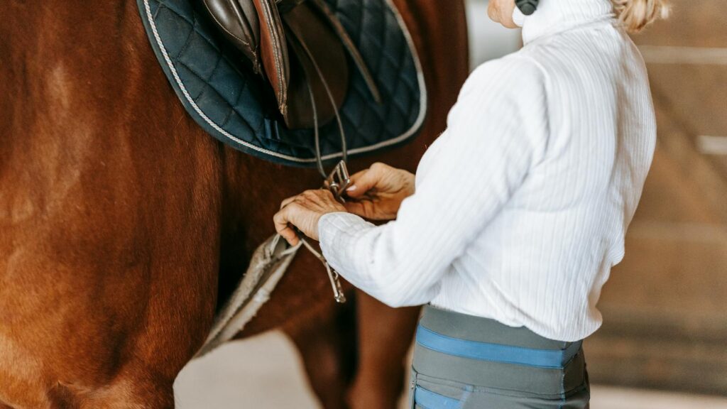 An equestrian woman adjusts a saddle on a horse inside a stable, preparing for a ride.