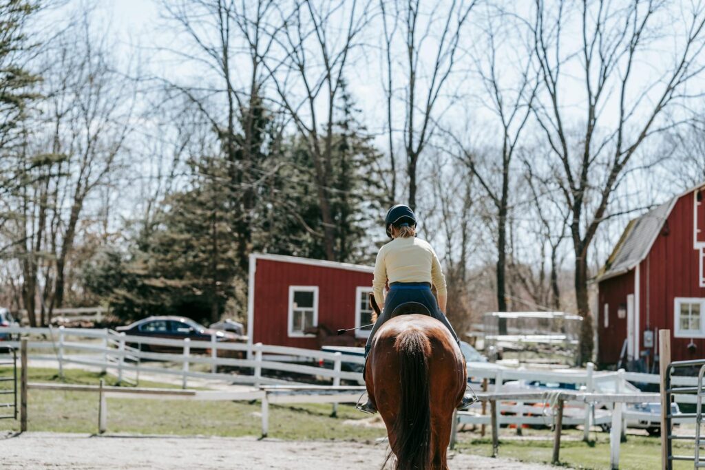 Back view of a girl horseback riding in a rustic paddock with red barns, enjoying leisure time outdoors.