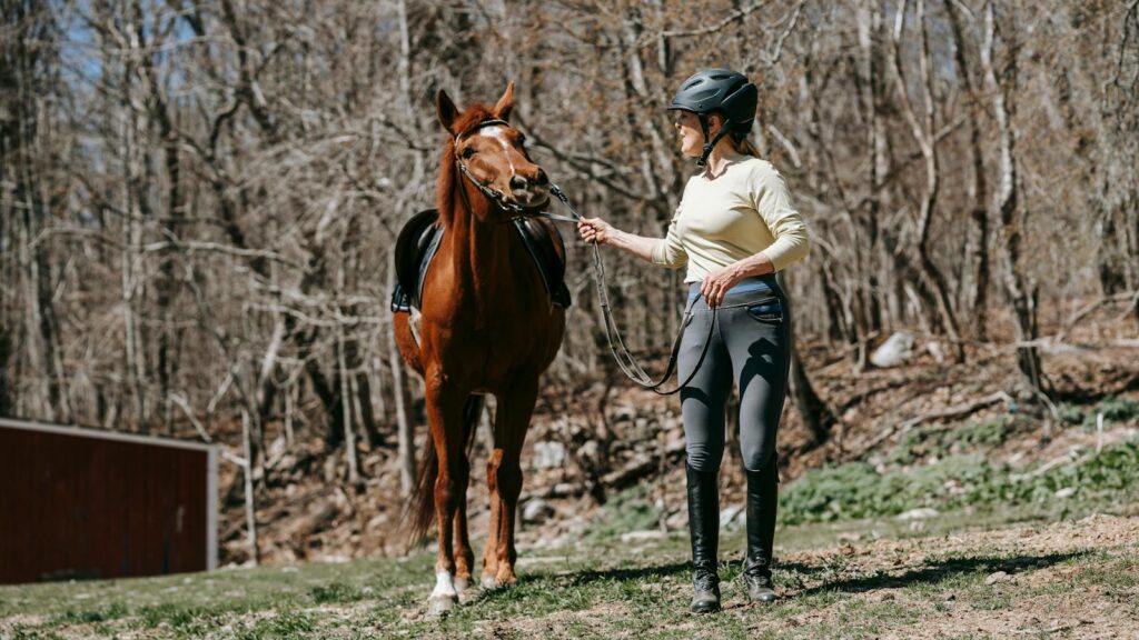 A woman leads a brown horse through outdoor training in a forest setting, showcasing equestrian skills.