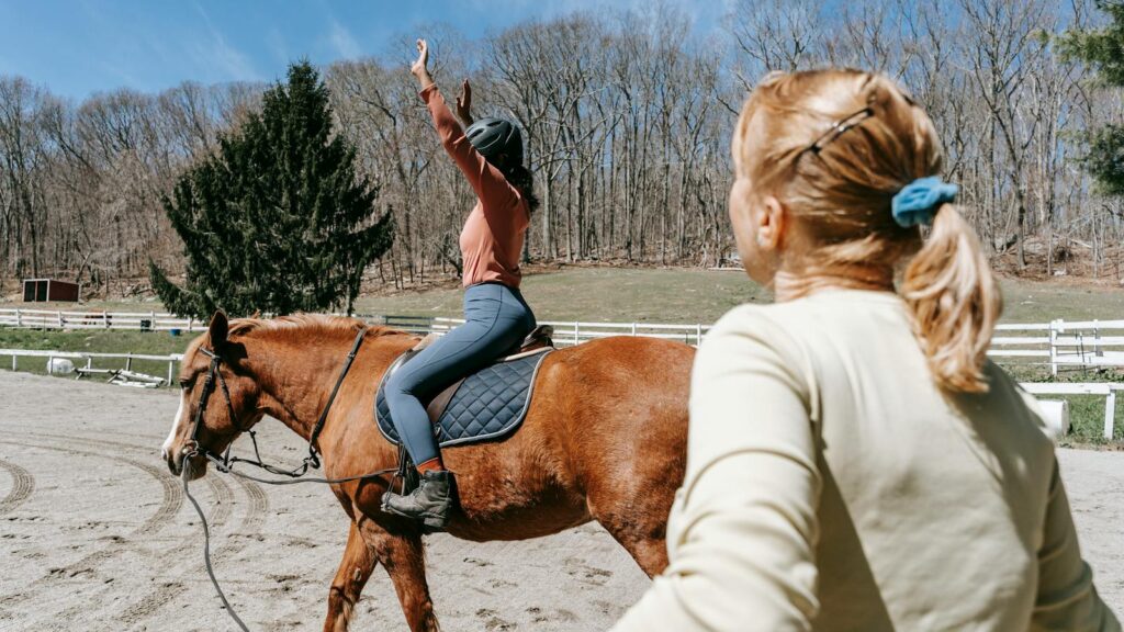 Woman learning horseback riding with an instructor in a sunny outdoor setting.