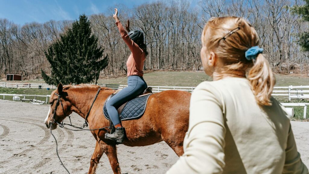 Woman learning horseback riding with an instructor in a sunny outdoor setting.
