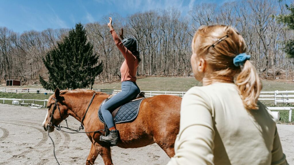 Woman learning horseback riding with an instructor in a sunny outdoor setting.