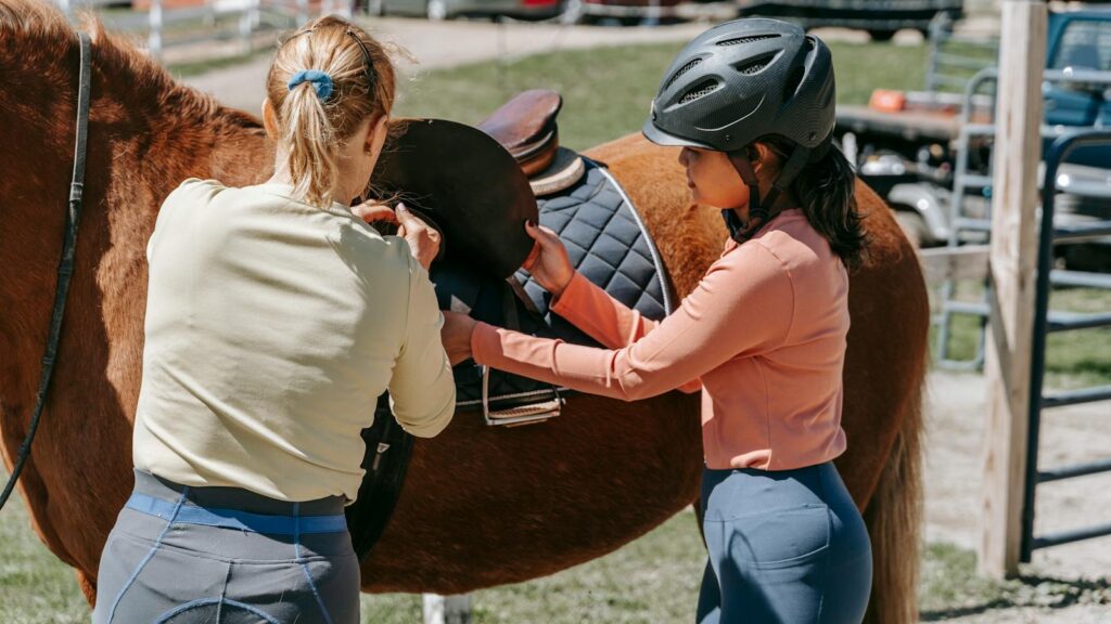 Two women preparing a horse for riding on a sunny day at a farm.