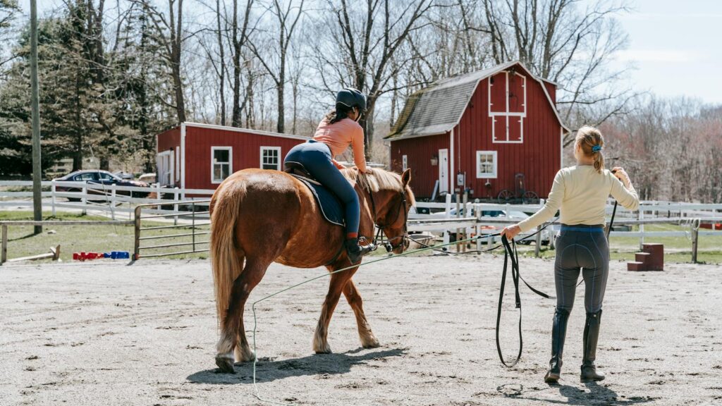A woman trains a horse rider outdoors near a red barn on a sunny day.