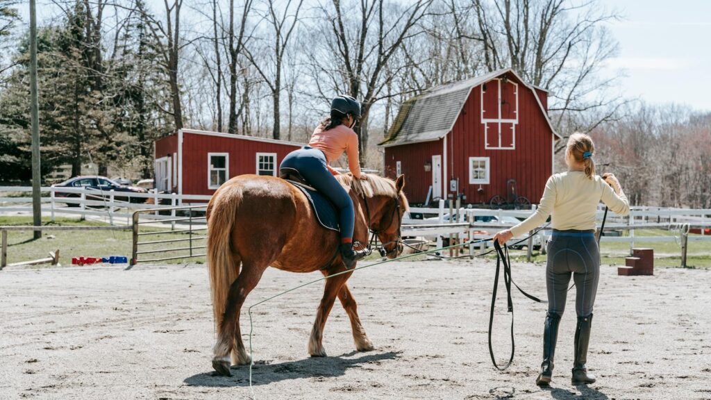 A woman trains a horse rider outdoors near a red barn on a sunny day.