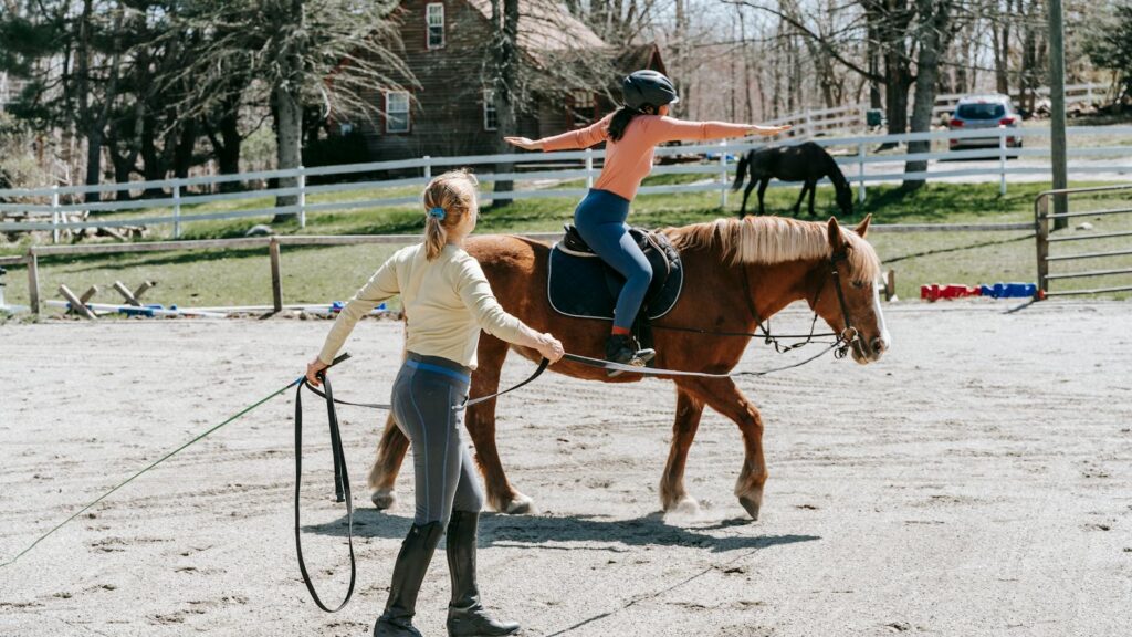 A young student learns horseback riding under the guidance of an instructor outdoors.
