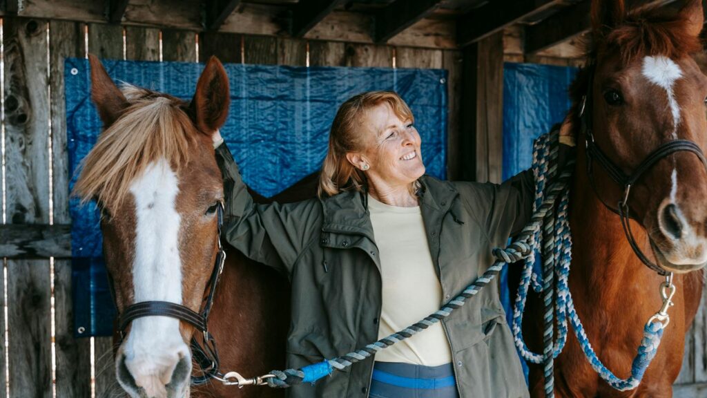 Smiling senior woman bonding with two horses in a rustic barn setting.