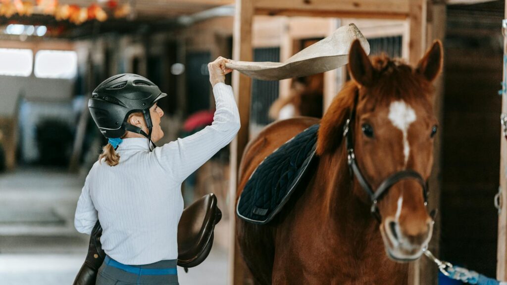 A woman in equestrian gear prepares a horse indoors in a stable setting.