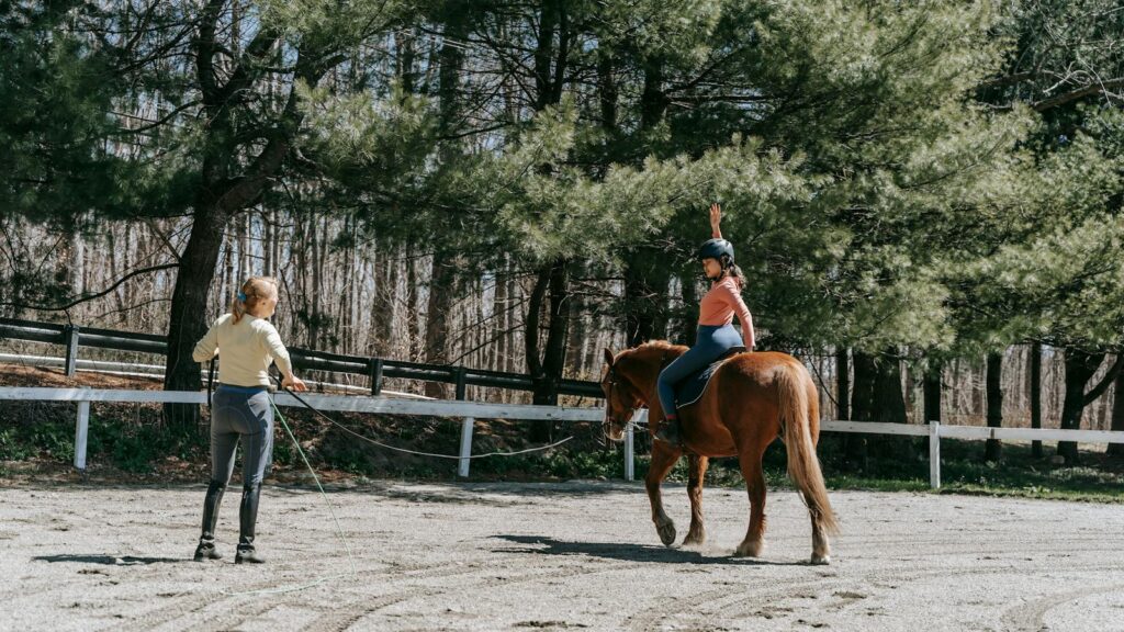 Woman instructing a child horseback riding under sunny forest trees.