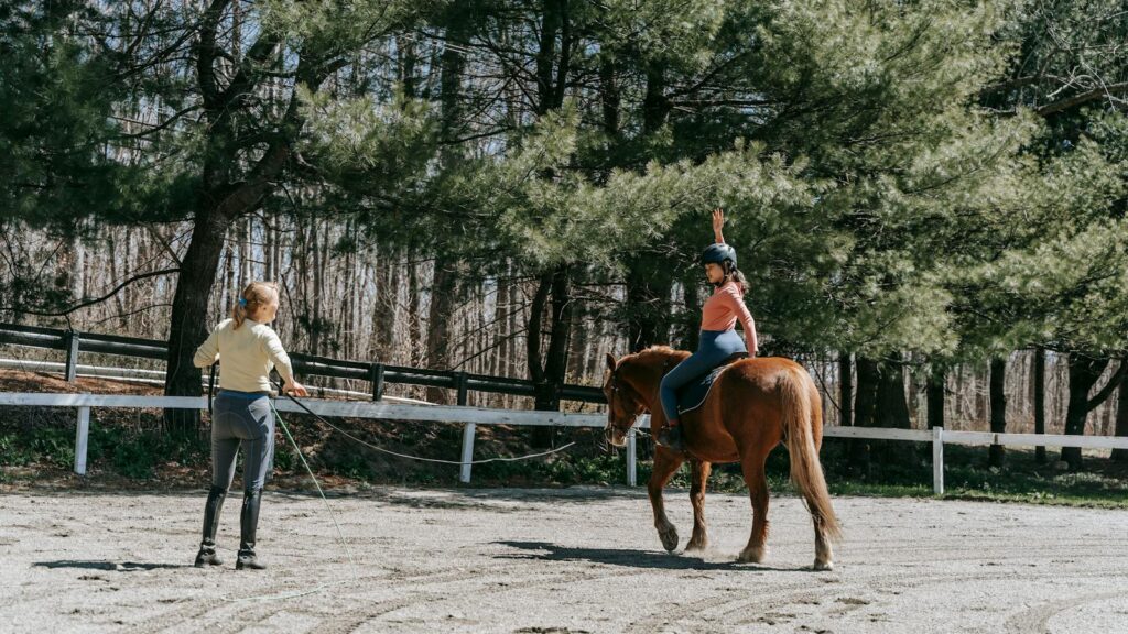 Woman instructing a child horseback riding under sunny forest trees.