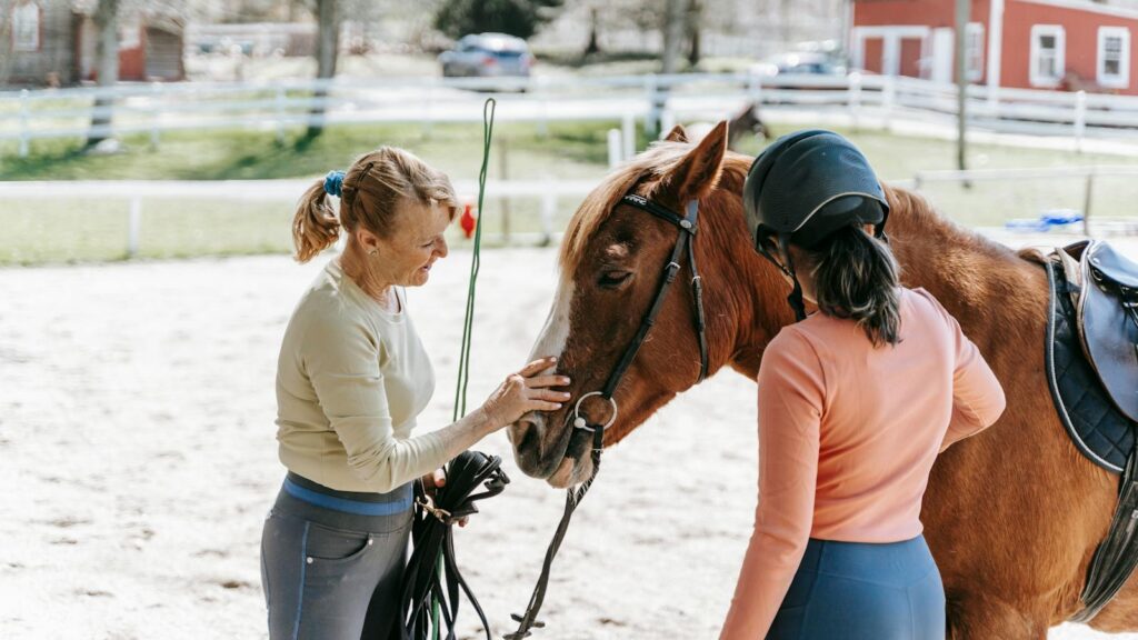Two women care for a horse at an outdoor stable, enjoying a sunny day.