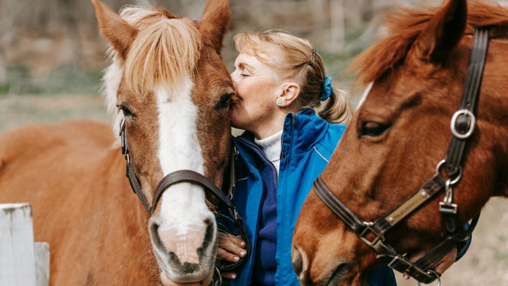 A woman lovingly kisses a horse on a sunny day, surrounded by nature.