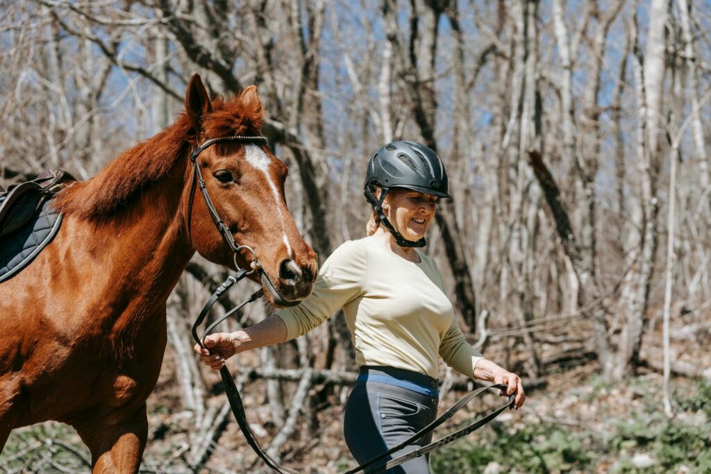A woman with a helmet leads a horse in a forest, enjoying an outdoor walk.