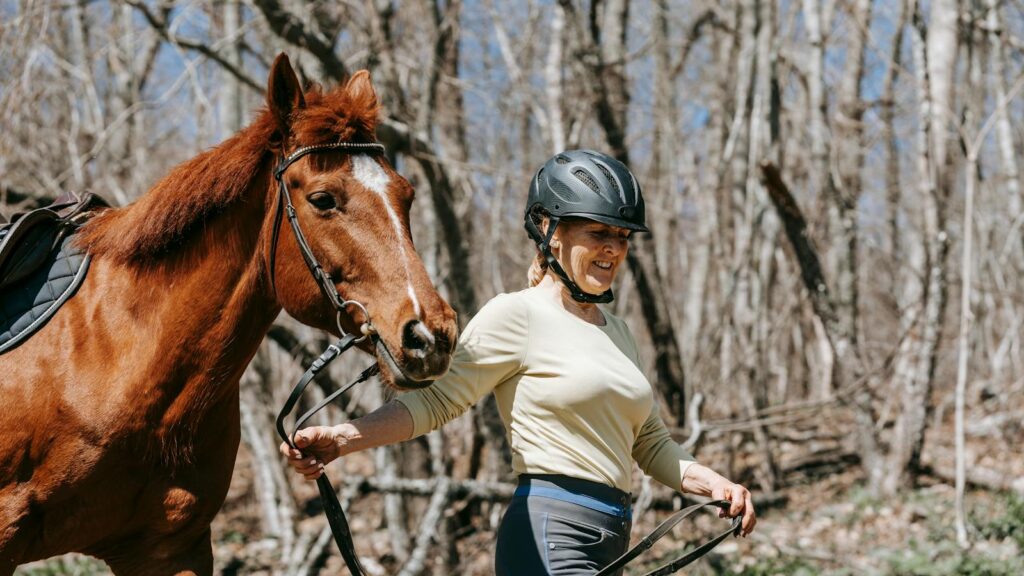 A woman with a helmet leads a horse in a forest, enjoying an outdoor walk.