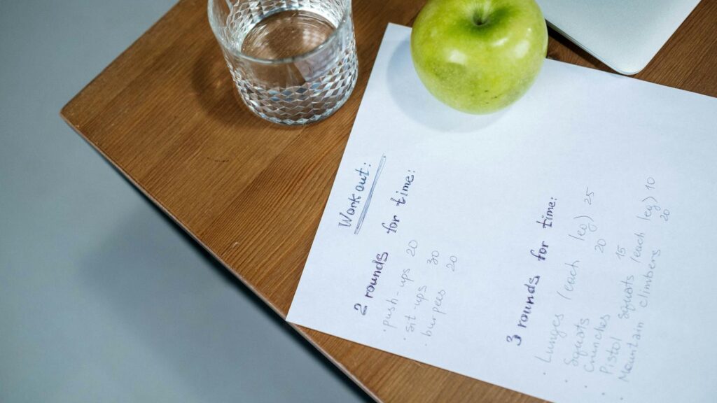 A green apple, glass of water, and workout plan on a wooden desk symbolize healthy living.