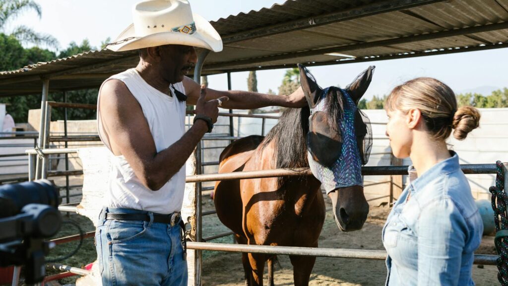A man in a cowboy hat and woman engage with a horse in an outdoor stable setting.