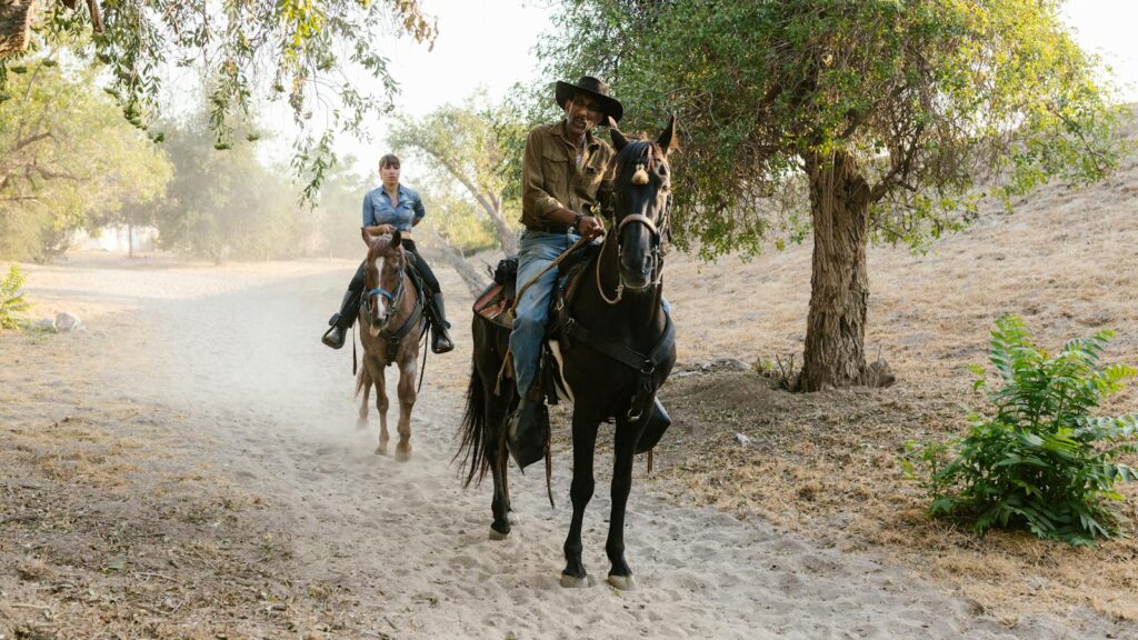 Two people riding horses on a rural dirt path surrounded by trees in a peaceful countryside setting.