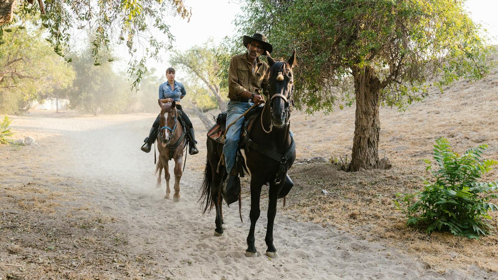 Two people riding horses on a rural dirt path surrounded by trees in a peaceful countryside setting.